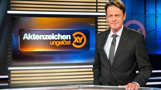 Ein Mann im Anzug steht in einem Fernsehstudio an einem Tisch.