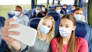 Zwei Mädchen sitzen mit Masken im Bus und machen ein Selfie.