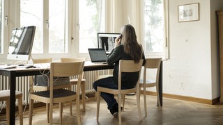 Eine Frau sitzt telefonierend an einem Tisch. Auf dem Tisch stehen zwei Computerbildschirme und ein Laptop.