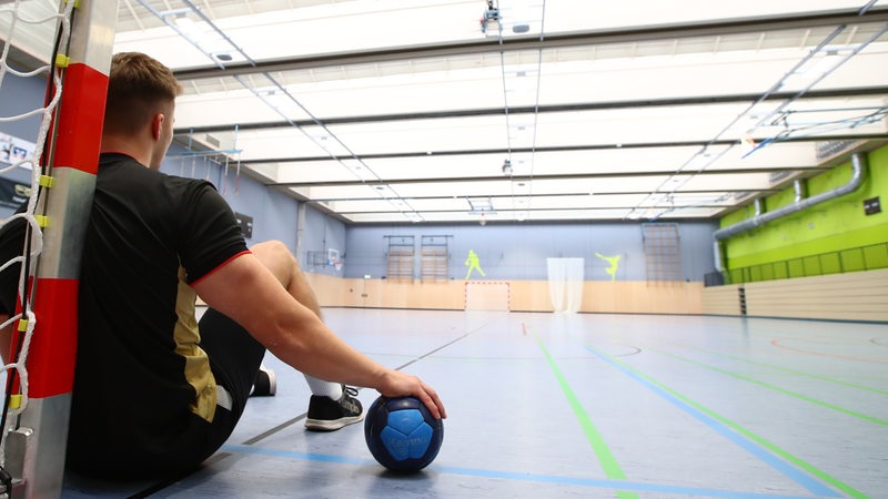 Ein Handballspieler lehnt in einer leeren Sporthalle mit einem Handball unter der Hand am Torpfosten.