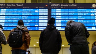 Menschen stehen in einem Bahnhof vor einer Anzeigetafel