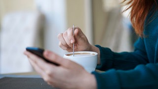 Eine Frau rührt einen Kaffee und schaut auf ein Smartphone (Symbolbild)