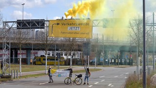 Aktivisten der Organisation "Extinction Rebellion" sitzen über einem Banner, das über einem Autobahnzubringer in Bremen hängt, und wedeln mit einer Rauchbombe. Mit der Besetzung der Traverse über der Fahrbahn wollen die Aktivisten auf ihre Belange im Zuge der in Bremen ausgerichteten Verkehrsministerkonferenz aufmerksam machen. 