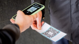Eine Person scannt vor einem Sicherheitsmitarbeiter mit der Luca-App einen QR-Code. 