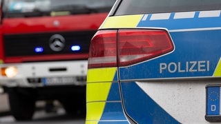 Ein Einsatzfahrzeug der Polizei mit Schriftzug, im Hintergrund ist ein Fahrzeug der Feuerwehr mit Blaulicht zu sehen. (Symbolbild)