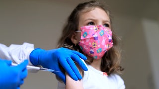 Ein kleines Mädchen mit rosa Mund-Nasen-Bedeckung bekommt eine Impfung in den Oberarm.