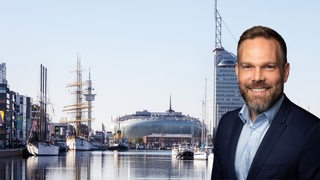Ein Mann mit Bart lächelt in die Kamera, dahinter der historische Hafen von Bremerhaven