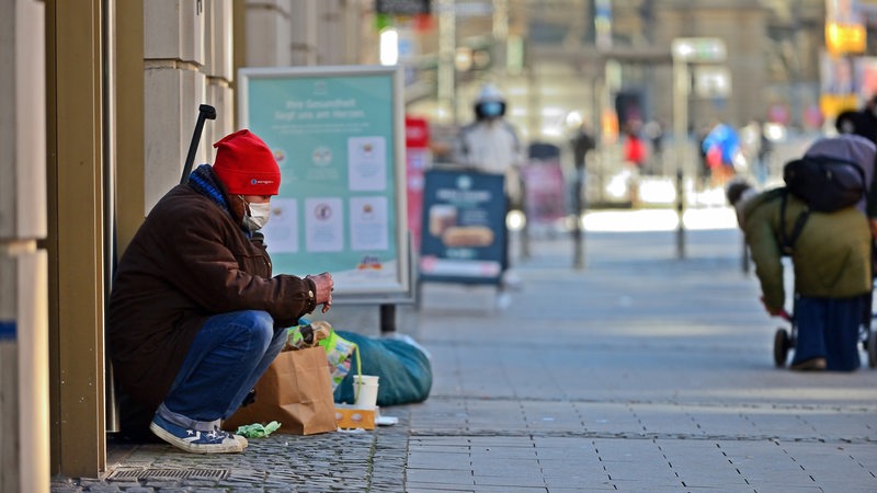 Ein Obdachloser sitzt an einer Hauswand in einer Einkaufsstraße.
