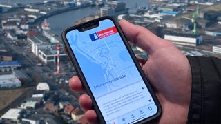 Eine Hand hält ein Smartphone mit dem Bremerhaven-Guide, dahinter der Fischereihafen (Montage)