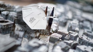 Collage zeigt eine FFP2-Maske, die an dem für Blinde gefertigte Bremen-Modell in der Innenstadt hängt