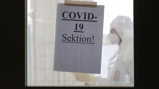 Rechtsmediziner steht hinter einer Tür eines Sektionssaals, an der ein Zettel mit der Aufschrift "COVID-19 Sektion!" (Symbolbild)
