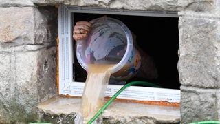 Ein Bewohner schüttet einen Eimer Wasser aus einem Kellerfenster nach draußen.