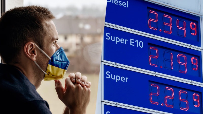 Ein Mann mit Schutzmaske in ukrainischen Farben, daneben Preistafel an einer Tankstelle (Montage)