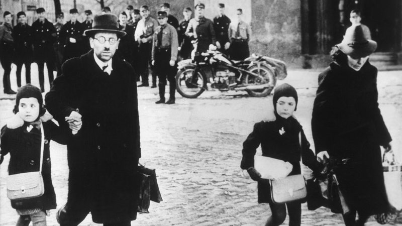 Eine jüdische Familie flüchtet mit wenig Gepäck unter dem höhnischen Blicken einer Gruppe von Nazis 1939