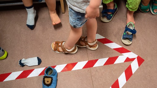 Kinder in einer Kindertagesstätte ziehen sich Schuhe an, auf dem Boden ein aufgeklebter Pfeil (Archivbild)
