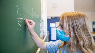 Eine Schülerin steht mit Schutzmaske vor einer Tafel und löst eine Rechenaufgabe (Archivbild)