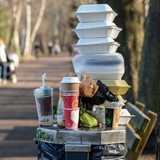 Auf einem vollen Mülleimer in einem Park stehen Kaffeebecher und Essensschachteln.