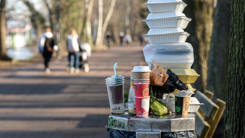 Auf einem vollen Mülleimer in einem Park stehen Kaffeebecher und Essensschachteln.