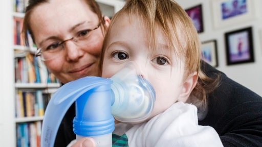 Ein zweijähriges Kind auf dem Arm der Mutter, dass an Mukovizidose erkrankt ist und inhaliert