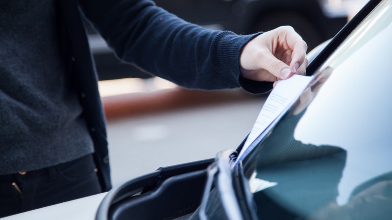 Eine junge Frau zieht einen Strafzettel unter ihrem Scheibenwischer hervor. 
