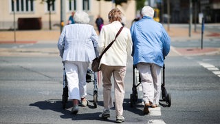 Drei ältere Damen - zwei von ihnen mit Rollatoren - gehen über eine Fußgängerampel. 