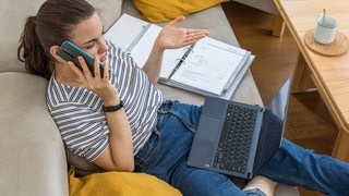 Eine Frau sitzt mit Laptop auf dem Schoß telefonierend im Homeoffice auf dem Sofa