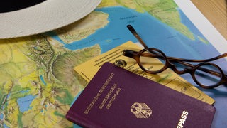 Reisepass, Impfausweis, sommerlicher Hut und eine Brille liegen auf einer Landkarte.