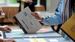 Ein Mann steckt seinen Stimmzettel in eine Wahlurne, ein Wahlhelfer schaut zu. (Archivbild)