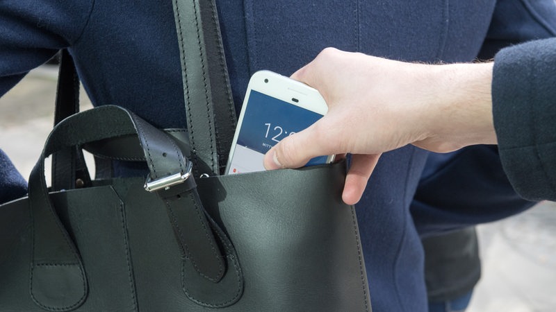 Eine Hand stiehlt ein Smartphone aus einer Handtasche.