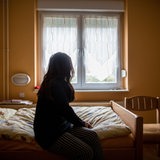 Eine Frau sitzt in einem Frauenhaus auf einem Bett und schaut zum Fenster (Archivbild)