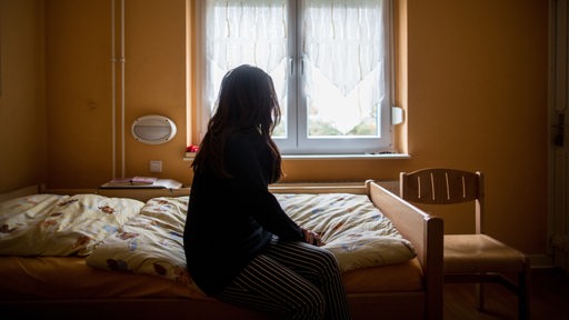 Eine Frau sitzt in einem Frauenhaus auf einem Bett und schaut zum Fenster (Archivbild)