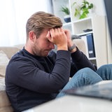 Ein junger Mann sitzt in einem Wohnzimmer und versenkt den Kopf in den Händen.