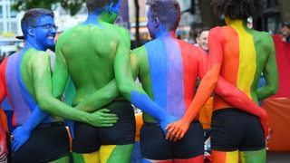 Vier junge Männer haben ihre Körper in Regenbogenfarben geschminkt und halten sich gegenseitig fest.