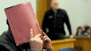 Ex-Krankenpfleger Niels Högel sitzt im Gerichtssaal und hält sich eine Akte vor das Gesicht.