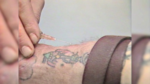 In einer Hand befindet sich eine Spritze, welche an einen Unterarm gehalten wird.