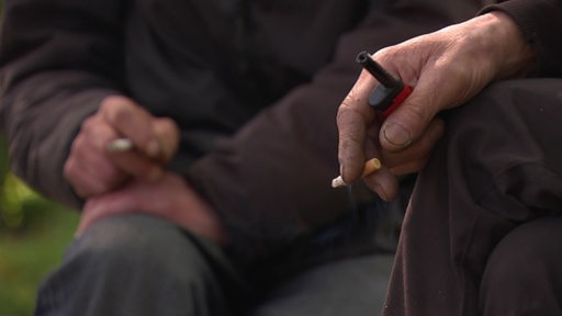 Im Vordergrund ist eine Hand zu sehen, welche zwischen den Fingern eine Zigarette und ein Feuerzeug hält. Im Hintergrund sitzt eine Person, die ebenfalls eine Zigarette zwischen den Fingern hält.