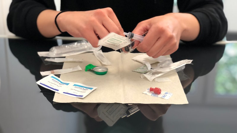 Eine Mitarbeiterin des Bremer Drogenkonsumraums zeigt an einem Tisch das typische Drogenbesteck, das Suchtkranke dort nutzen können.