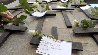 Eine weiße Rose befestigt eine Frau an einer Wand mit symbolischen Grabkreuzen anlässlich des Gedenktages für verstorbene DrochgebraucherInnen" 