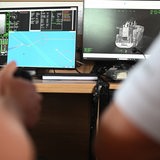 Zwei Personen sitzen mit einem Steuerknüppel vor zwei Bildschirmen worauf ein Schiff zu sehen ist.