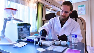 Ein Mann in einem weißen Kittel zeigt ein Testverfahren für das Drug Checking in einem mobilen Labor.
