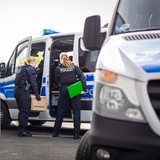 Eine Polizeibeamtin steht während einer Durchsuchung mit einem Karton an einem Polizeiwagen.