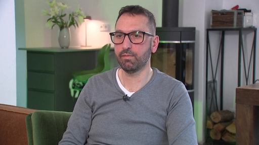 Ein Mann sitzt auf einem Stuhl, er trägt eine eckige Brille und einen grauen Pullover.
