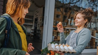 Eine junge Frau eines Gemüseladens hält eine Eierpalette im Arm und bietet einer Käufern ein braunes Ei an.
