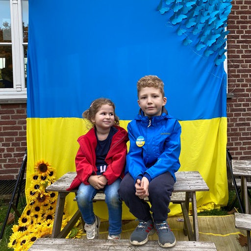 Zwei Kinder sitzen vor einer Ukraine-Flagge auf einer Bank