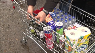 Ein Einkaufswagen befüllt mit Lebensmitteln. Es befinden sich Weinflaschen, Chips, Küchenrolle und andere Dinge in dem Wagen. Zwei Hände greifen nach den Weinflaschen.