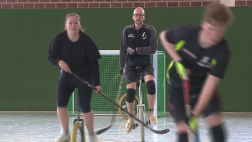 Es sind drei EinradhockeyspielerInnen in einer Sporthalle zu sehen. In der Mitte fährt der Nationalspieler Malte Voekel auf seinem Einrad.