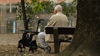 Ein alter Mann sitzt alleine auf einer Parkbank. Neben ihm steht sein Rollator