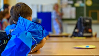 Ein Kind sitzt mit blauer Schultüte im Klassenzimmer und schaut zur Lehrerin an der Tafel.