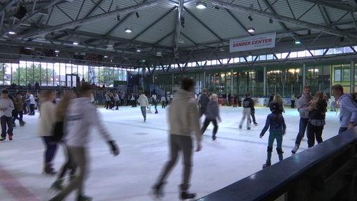 In einer Eislaufhalle sind mehrere Personen mit Schlittschuhen auf der Eisbahn zu sehen.