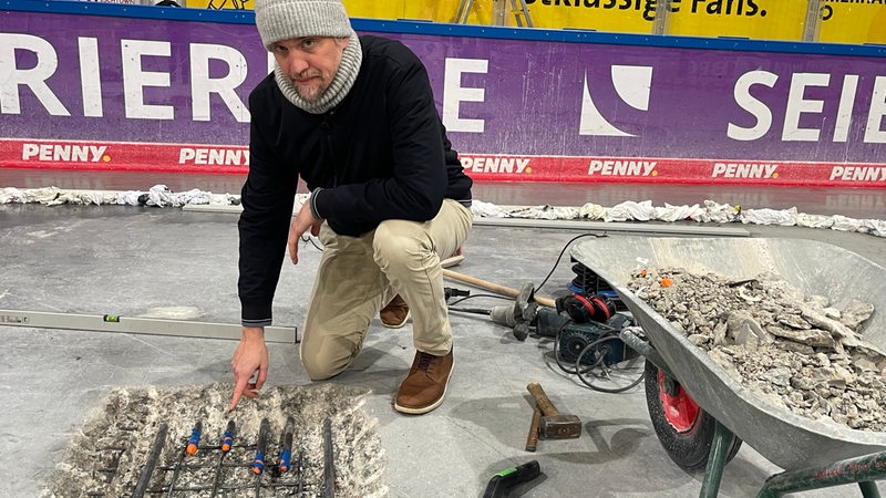 Stadthallenchef Gimpel zeigt auf ein Loch im Boden der Eisarena Bremerhaven. Neben ihm steht eine Schubkarre.